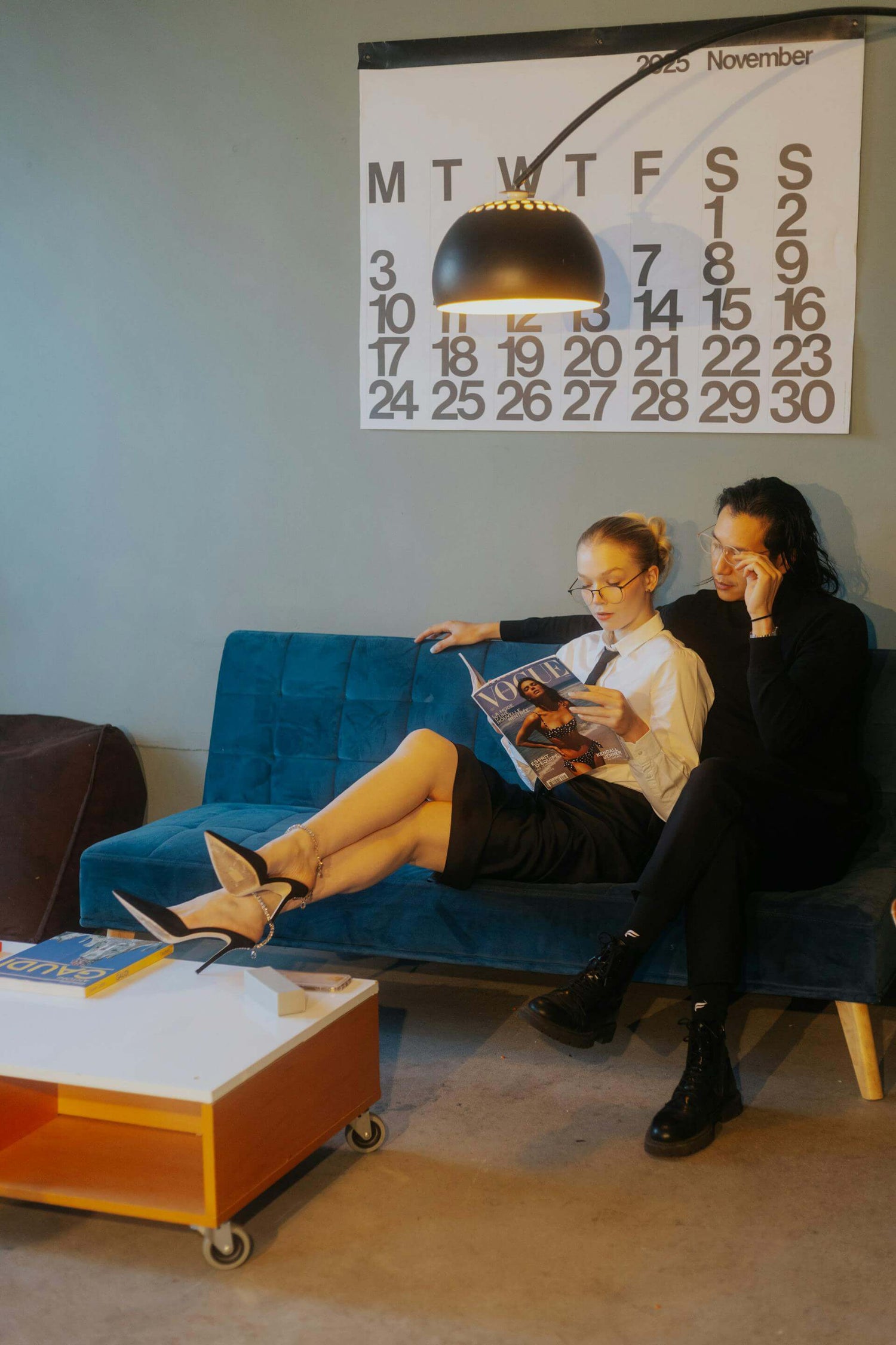 Small space urban living room with two people sitting on a blue couch in a room with a calendar on the wall.